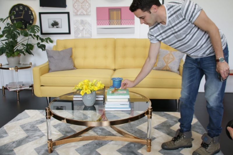 Kyle decorating glass cocktail table in front of yellow sofa