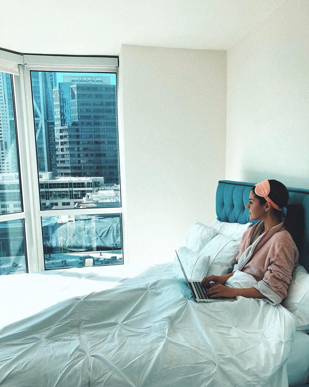 woman in blue bed in pink robe on laptop looking out the window 