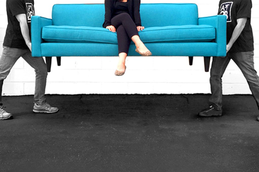 two men lifting up a blue sofa with a woman sitting on it