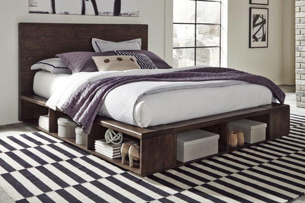 room with wooden bed, purple blanket, pillows with storage underneath bed placed on a black and white striped rug