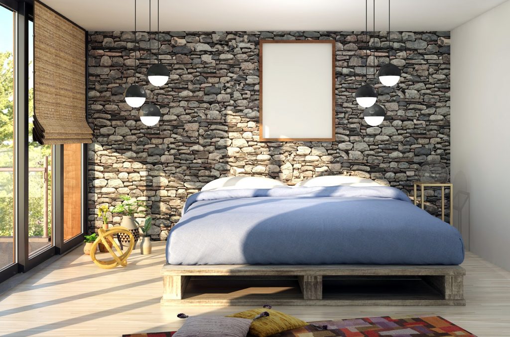 Grey wooden slat bed with blue sheets in front of a stacked stone wall with two hanging lights