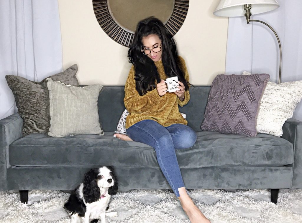 woman on grey sofa drinking out of a mug with a dog on the rug