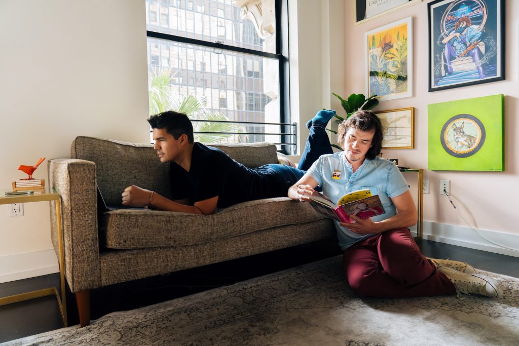 two men in a room with a grey sofa hanging out with a book and paintings