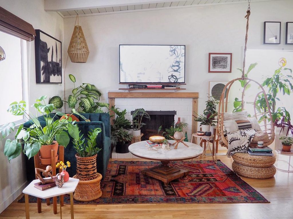 room with a ton of plants, a red rug, green sofa, tv, wall art, paintings, white coffee table, books, baskets and wooden chairs