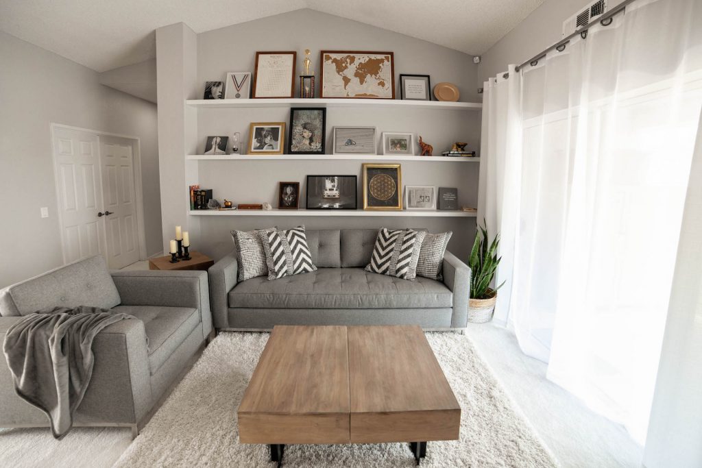 room with two grey sofas, wooden table, beige rug, paintings and pictures, candles and a large window with a curtain