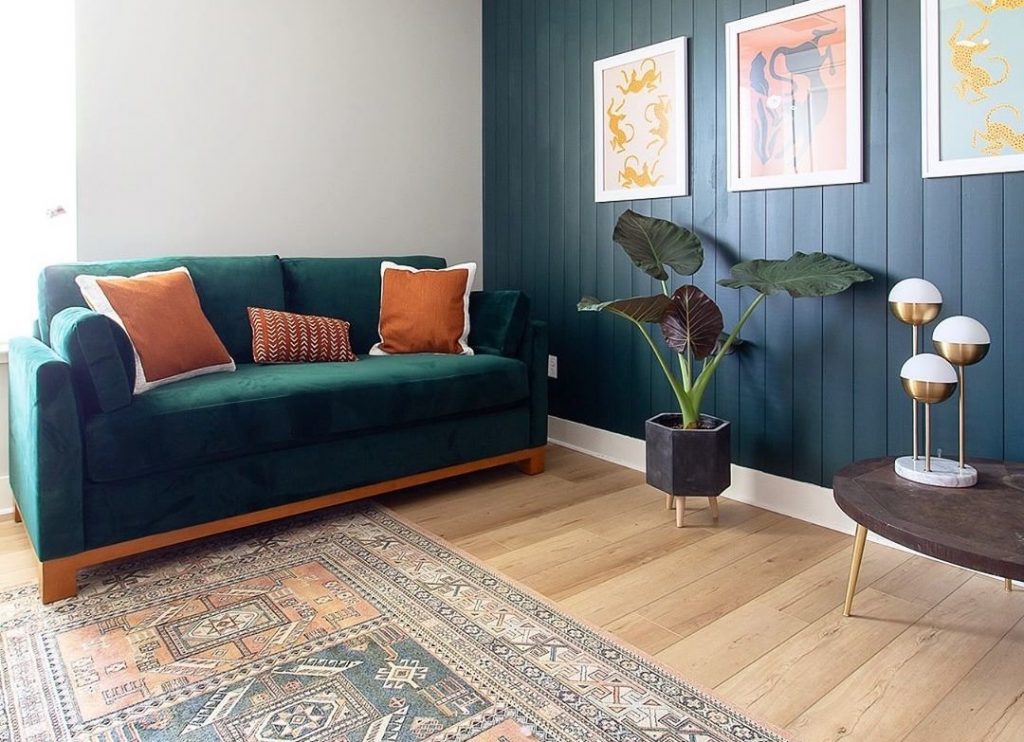 green sofa with orange pillows, a plant, wooden table, lamp, paintings, and rug