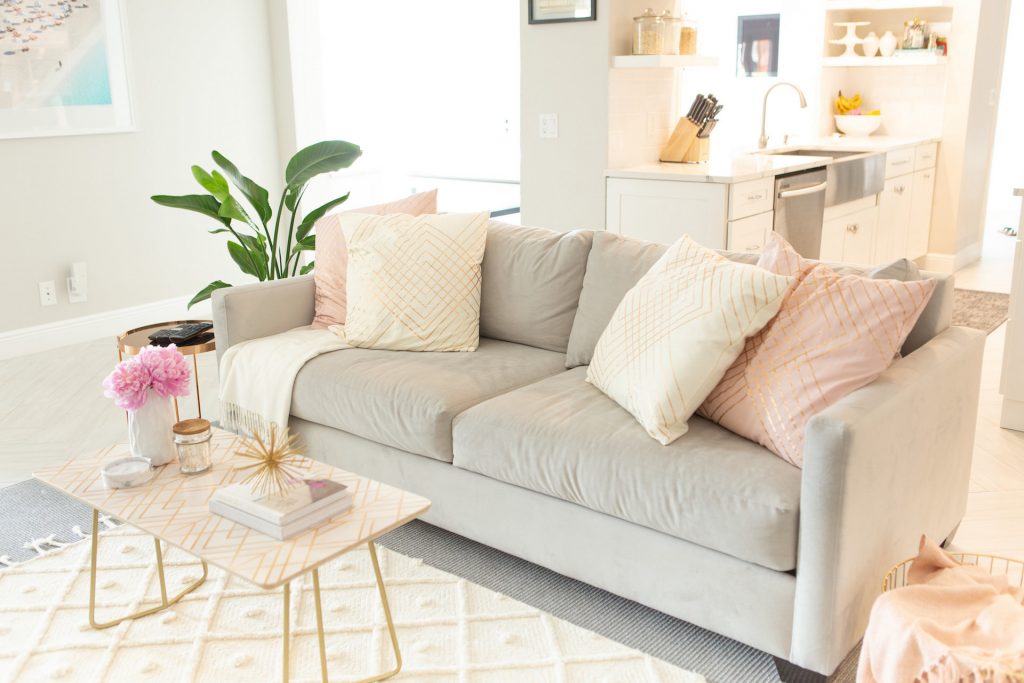 room with a grey sofa, pink and white pillows, purple ottoman, plant, gold side table, white table, paintings and white rug