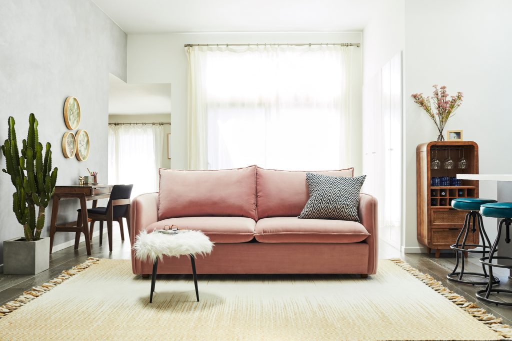 pink sofa with grey pillow, beige rug, fuzzy white ottoman, cacti, brown desk with chair and wall clocks