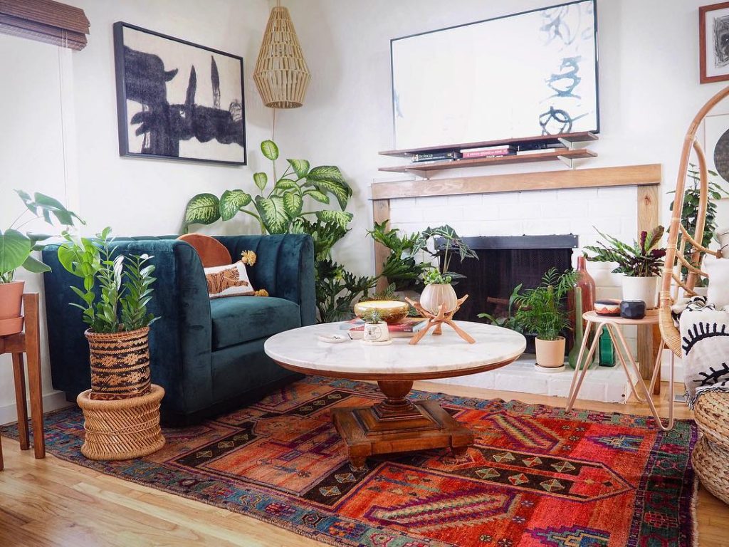 green chair, white table, red rug, baskets, multiple plants facing a tv, decorative ceiling baskets and paintings
