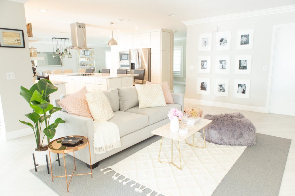 room with a grey sofa, pink and white pillows, purple ottoman, plant, gold side table, white table, paintings and white rug