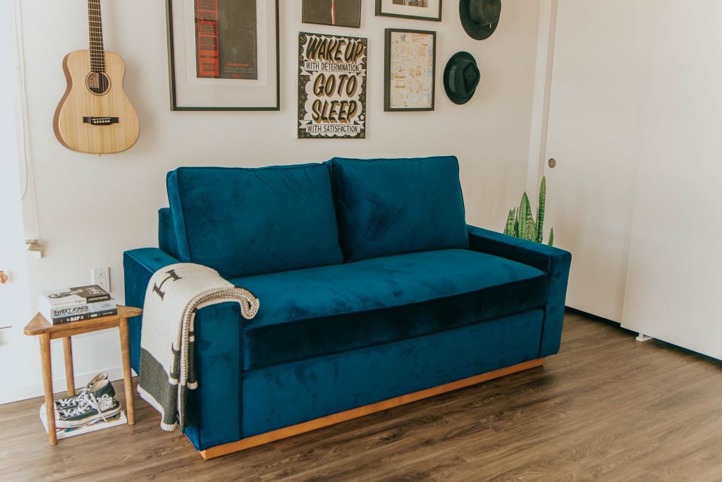 blue sofa with blanket on it with a plant, side table, books, paintings and a guitar surrounding it