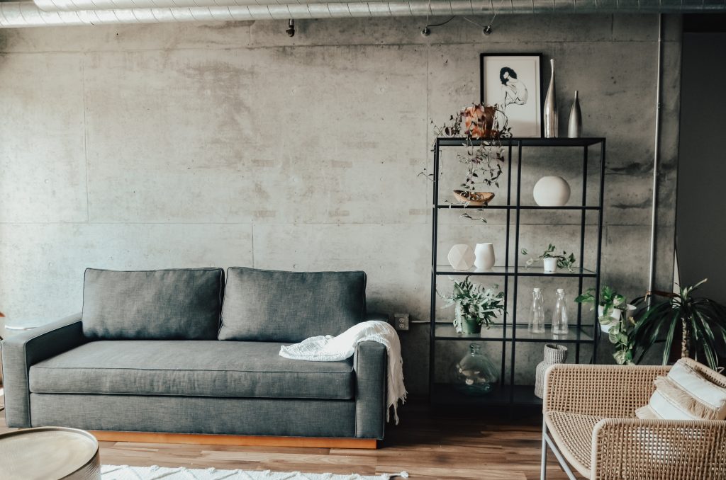 grey sofa with a blanket on it beside a book shelf, beige chair, ottoman and painting