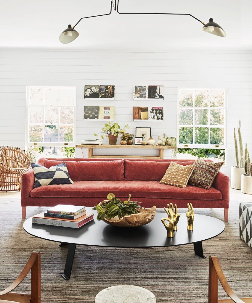 red sofa with pillows on it next to a black round coffee table, brown chairs, ottoman, plants, paintings and table with plants on it