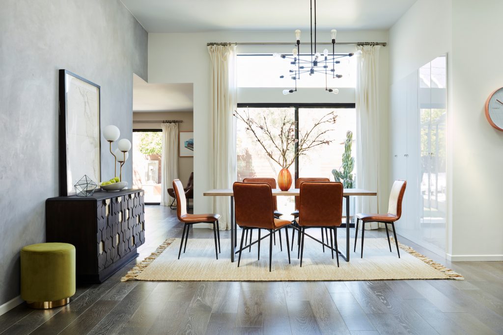 room with orange chairs, a dining table, large vase with plant, lamp, green ottoman, black cabinet, beige rug and large window with curtains