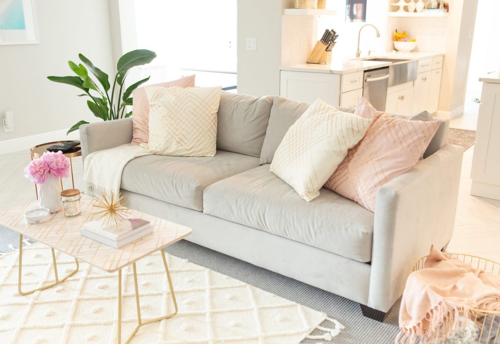 beige sofa with pink and white pillows beside a pink and gold coffee table, plant, gold side table, a basket with a pink blanket all set on a grey and white rug