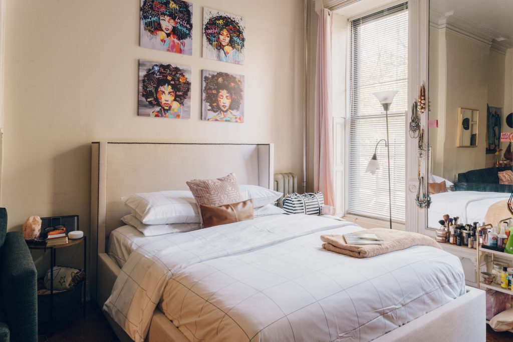 beige bed with different colored pillows surrounded by paintings, a black side table, lamp and beauty supplies