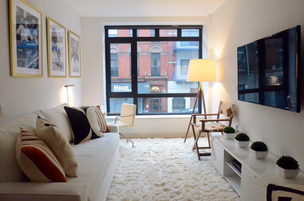 room with white sofa and different colored pillows placed on a white furry rug with a white chair, lamp, brown and white chair, white console, tv and plants