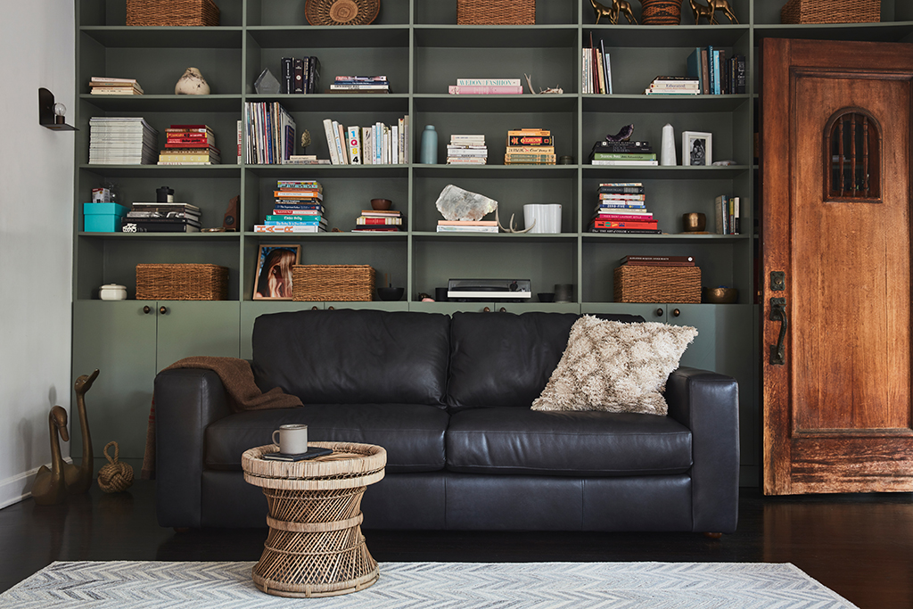 black leather sofa with pillow and blanket on it beside a book shelf and wooden ottoman