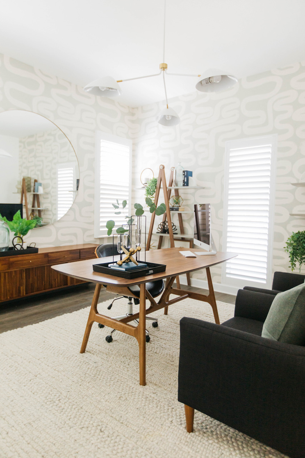 room with two black chairs with pillows, a wooden table with art pieces on them, a mirror, a book shelf, cabinet, plants and a beige rug