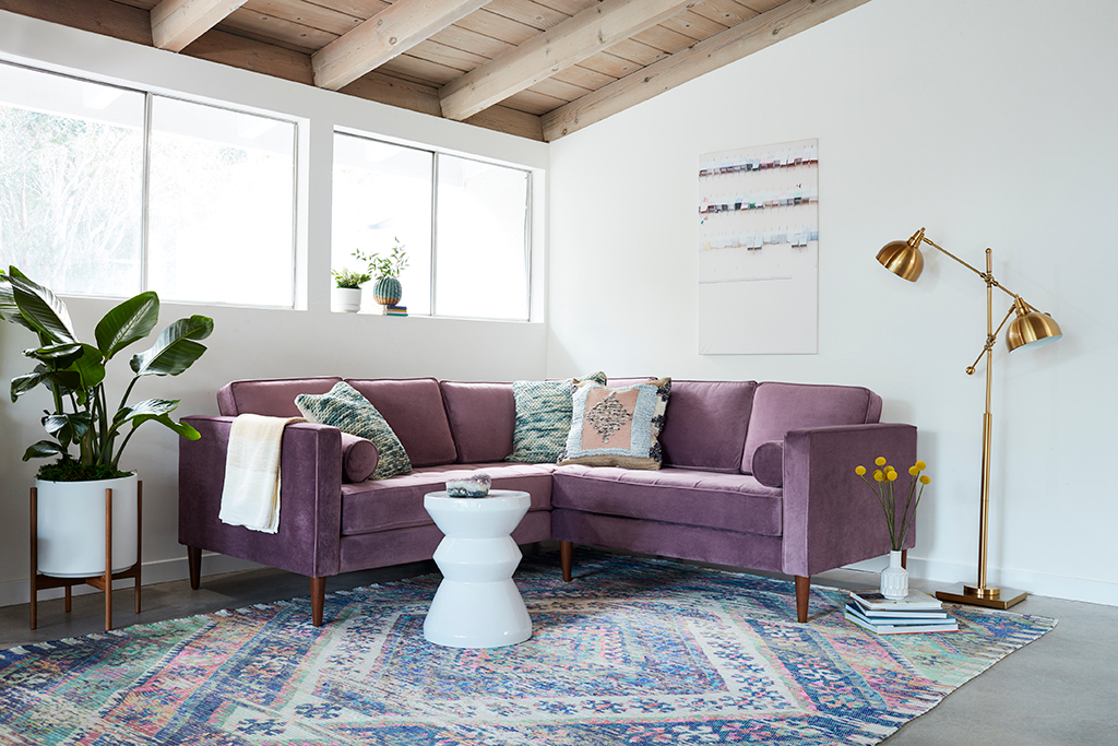 purple sofa with pillows next to a white coffee table, plant rug and lamp