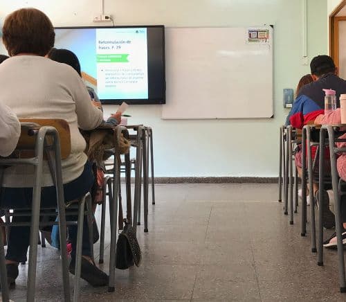 Persona reunidas en un salón de clases, mientras reciben alguna clase que ven un televisor