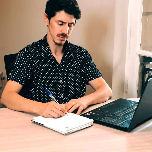 Hombre joven escribe algo en un cuaderno con el lapicero que sostiene con sus dedos, este está frente al computador