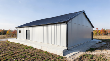 A modern light-gray steel workshop building with a charcoal gray roof, built on a clean concrete foundation in a rural landscape.