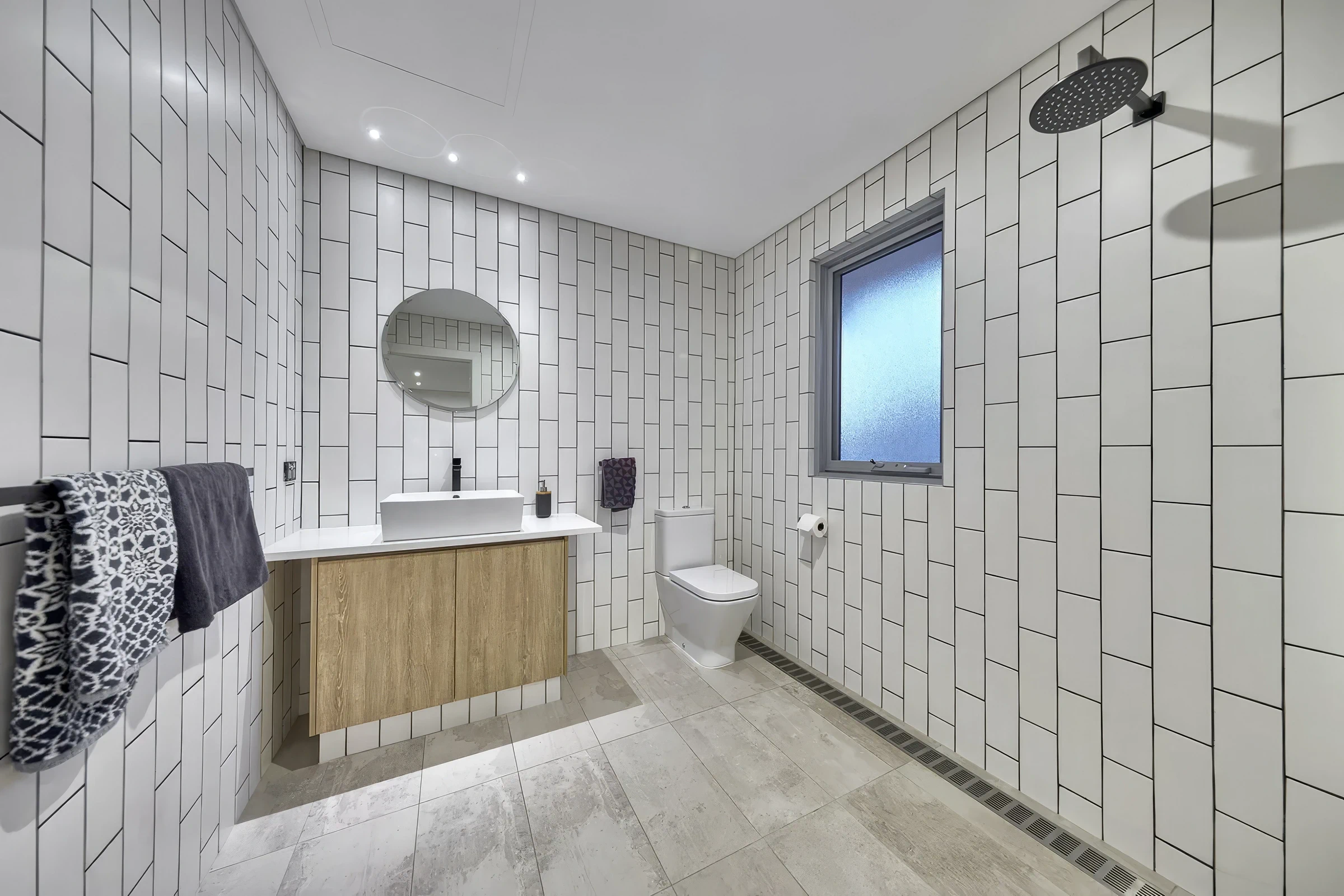 Bathroom with white tiled walls, wooden vanity, round mirror, toilet, frosted window, and rainfall shower head