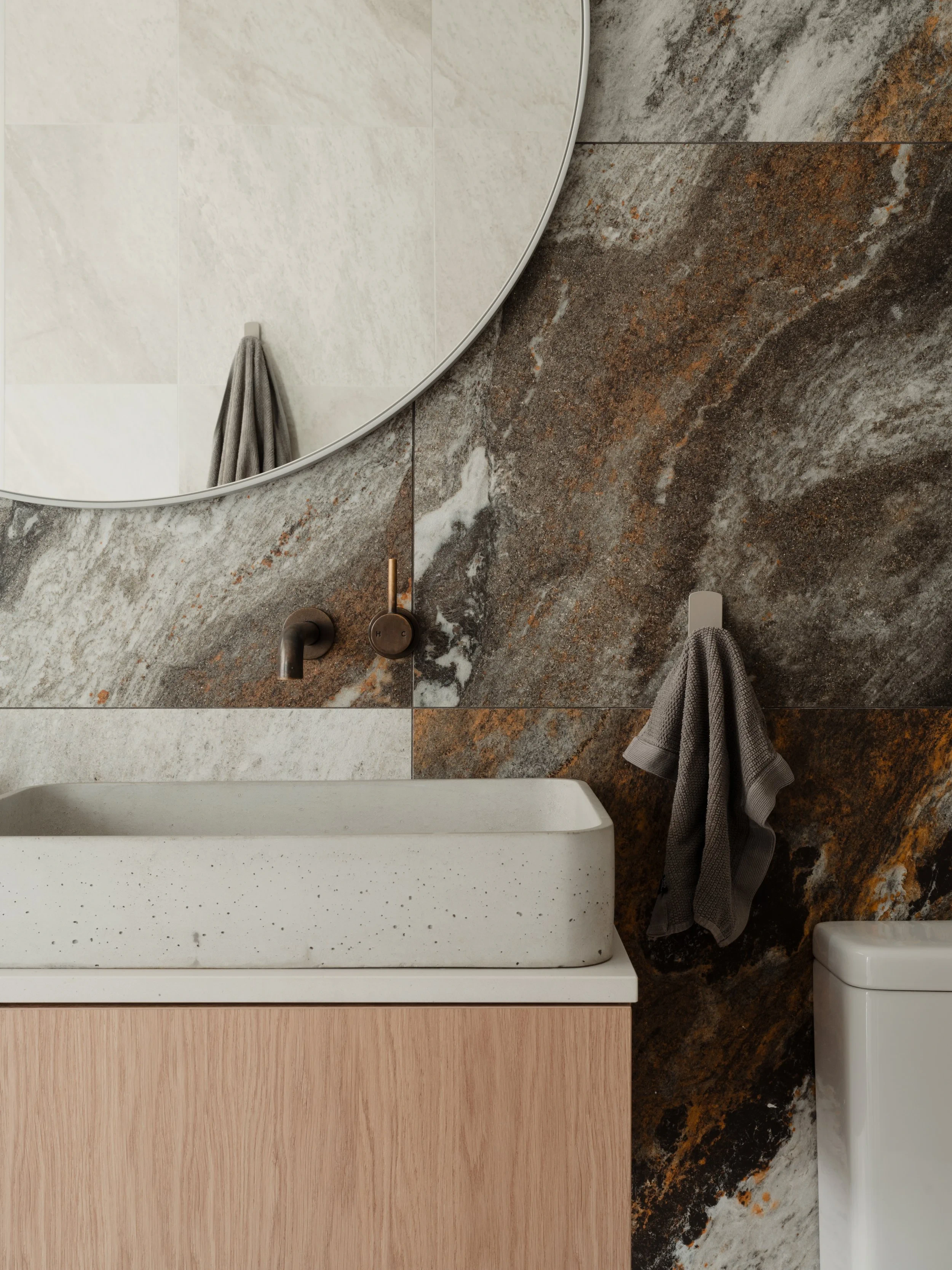 Bathroom with a large round mirror, textured dark wall tiles, a white countertop basin, and wall-mounted faucet, with a grey towel hanging on a hook.