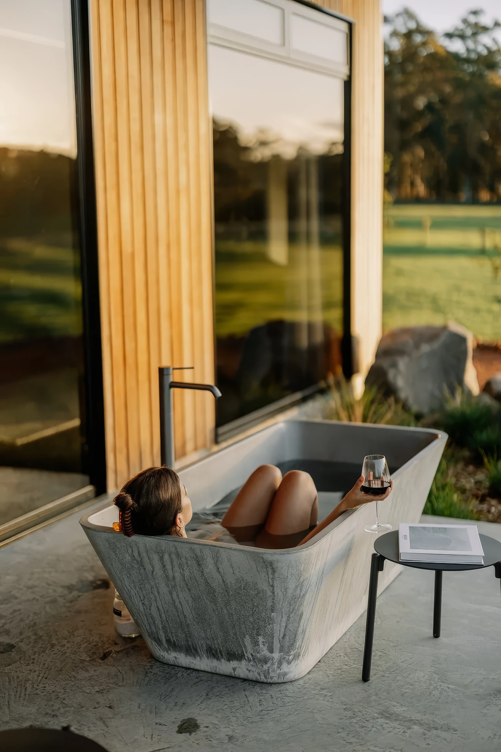 Woman relaxing in a freestanding bathtub outside, holding a glass of red wine