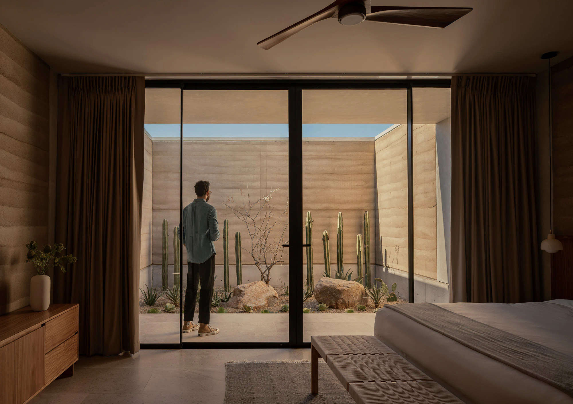 Indoor bedroom with a view of a small desert garden featuring tall cacti and rocks through large sliding glass doors