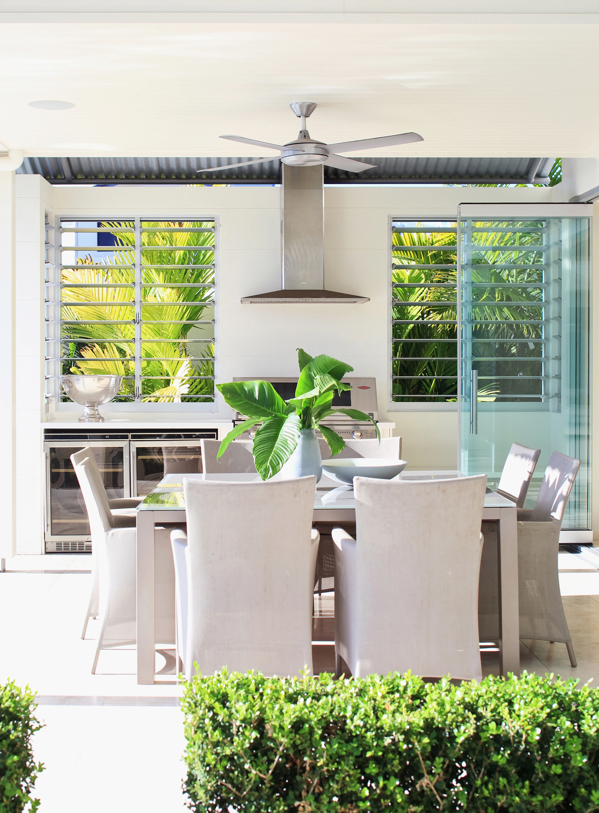 Outdoor dining area with a white table, six white chairs, potted plant, louvered windows, stainless steel range hood, ceiling fan, glass doors, and tropical greenery in a modern home setting