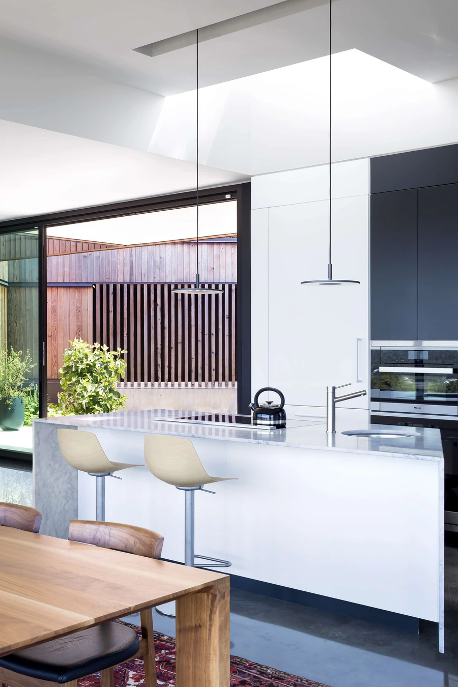 Modern minimalist kitchen with white island, bar stools, and view of outdoor wooden enclosure