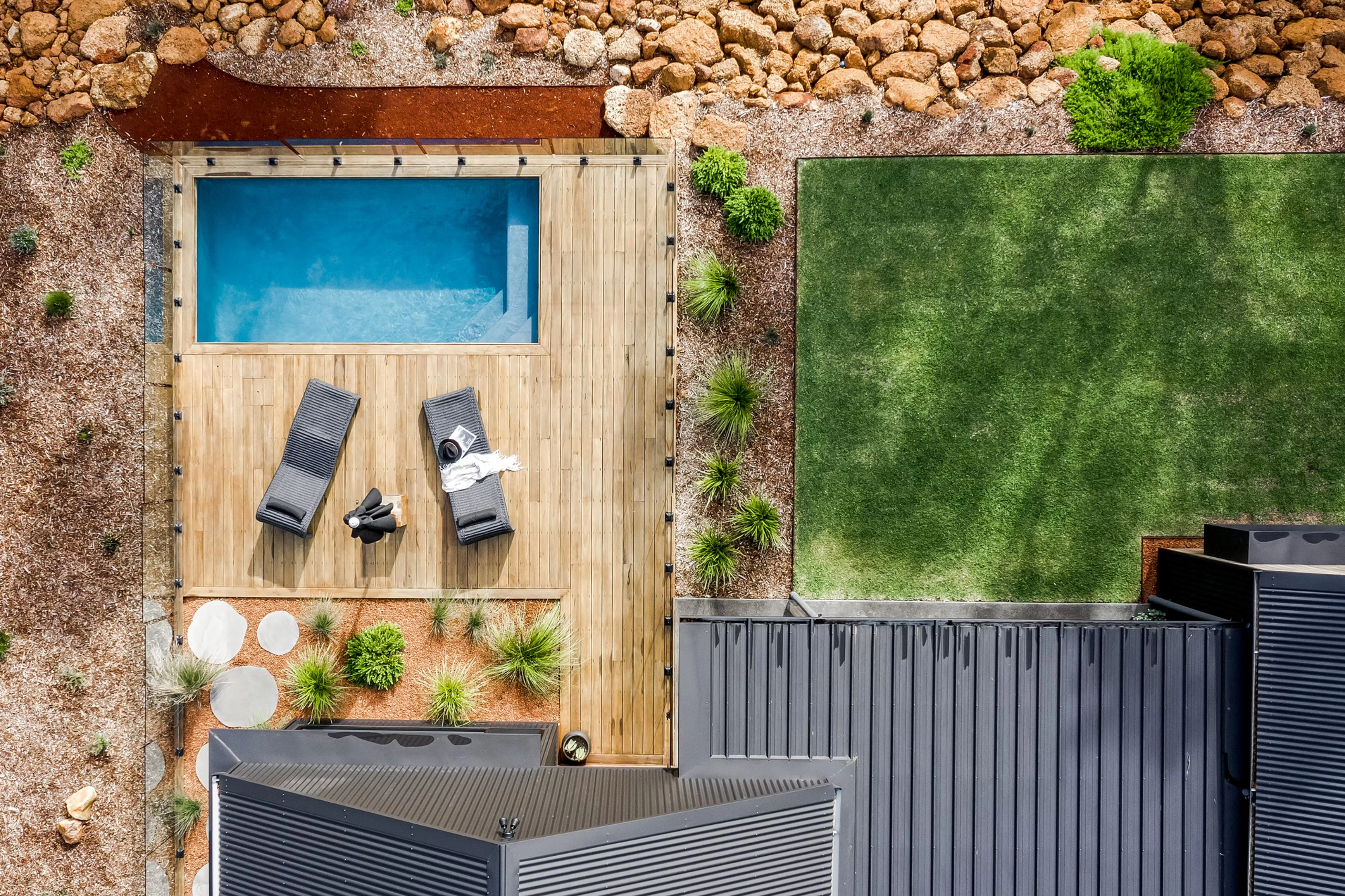 Aerial view of a backyard with a swimming pool, wooden deck, garden grass, and surrounding landscaping