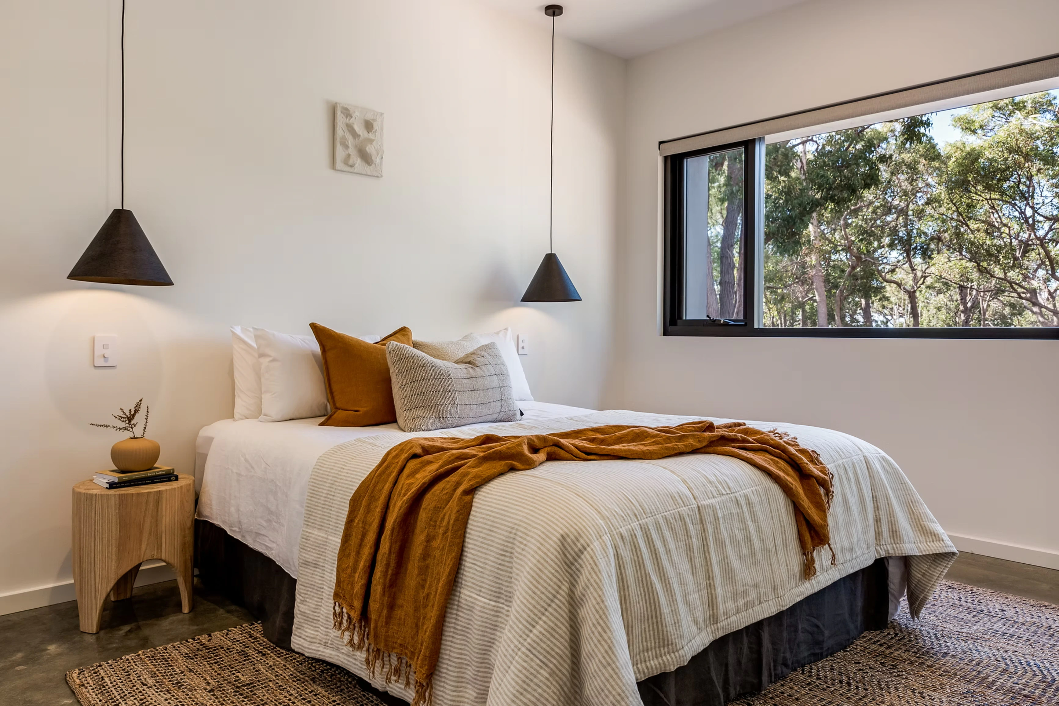 Minimalist bedroom with a large window showing greenery, white walls, beige bedding, and decorative cushions