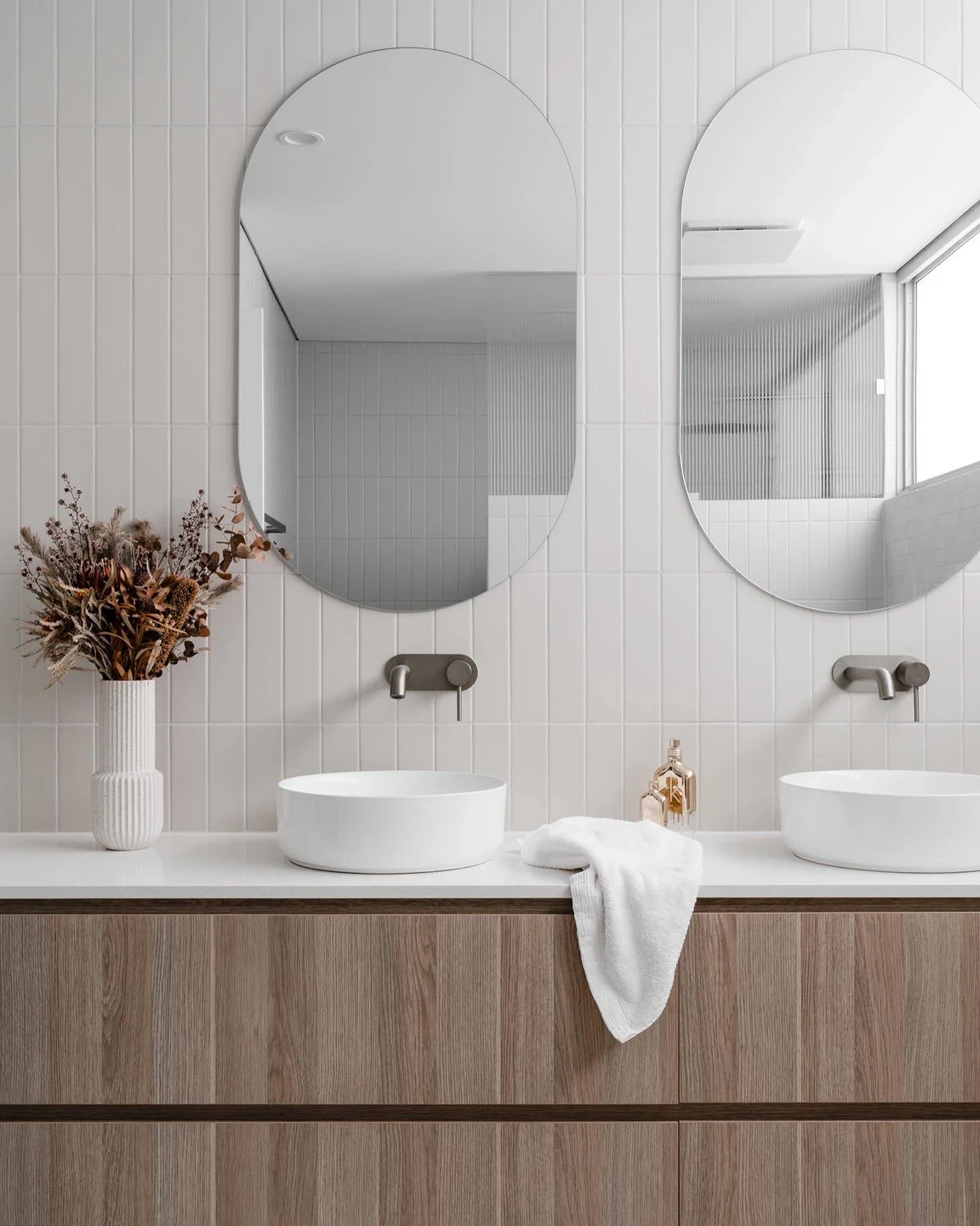 Bathroom vanity with two oval mirrors, vessel sinks, wooden cabinet, dried flower arrangement, towel, and decorative soap containers