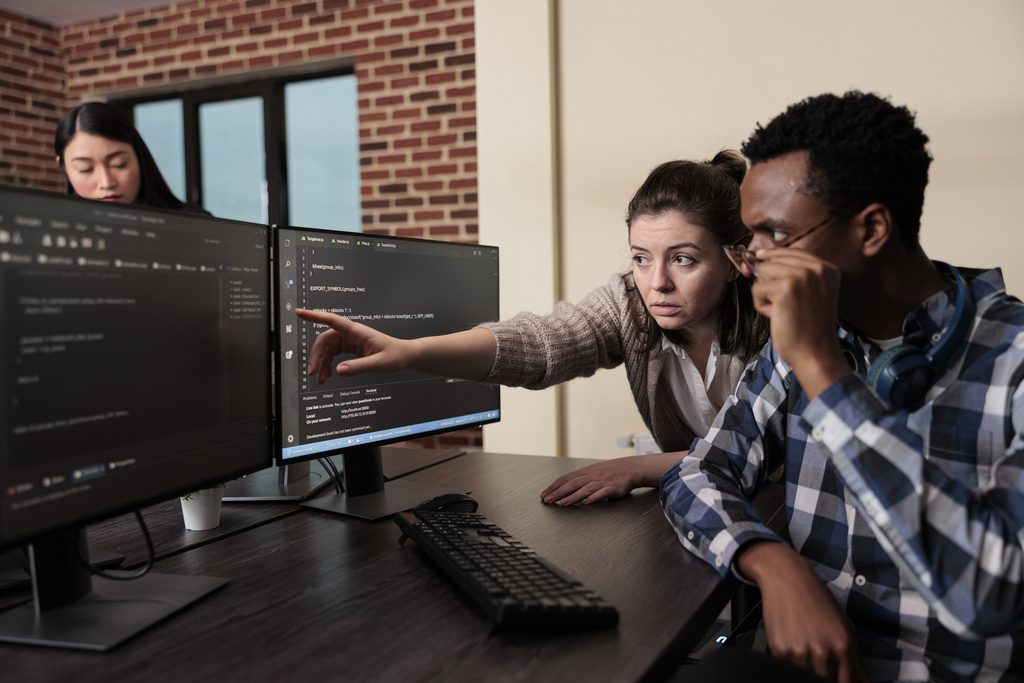 Mulher branca e homem negro olham para o computador enquanto ela aponta para uma linha de código, servindo como ilustração para a temática do texto de revisão de código.