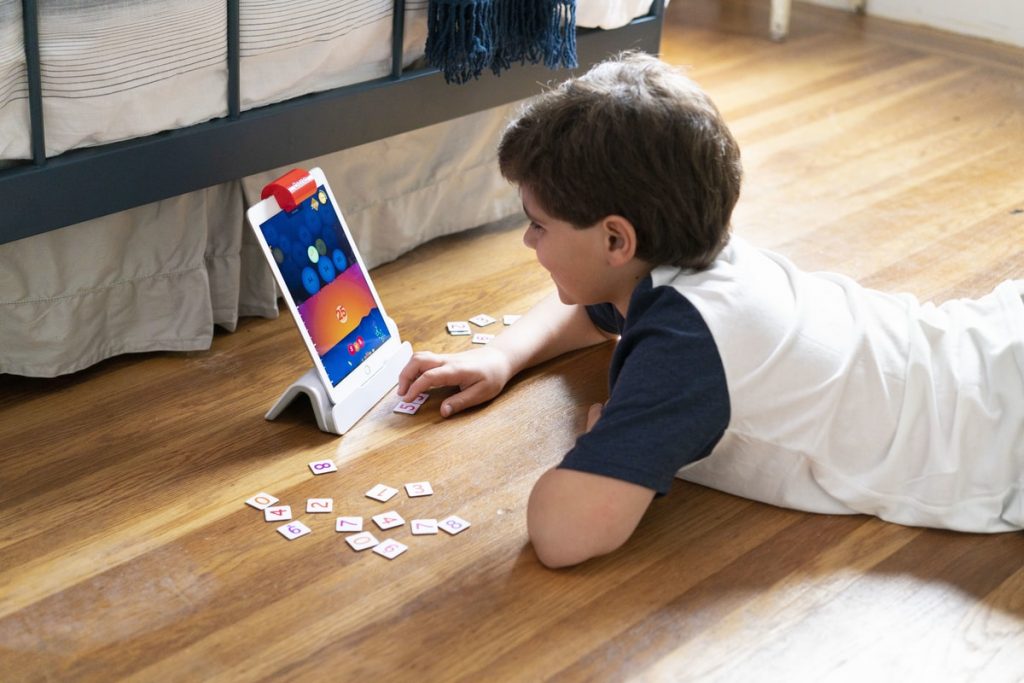  A boy is lying on the floor and using Osmo, an interactive learning game that uses a tablet and physical tiles to teach math, coding, and other subjects.