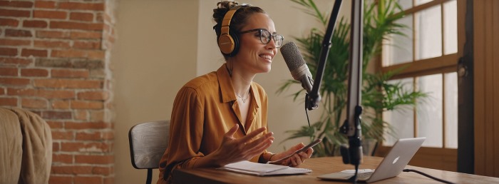 Woman wearing headphones and speaking into a studio microphone at a desk with laptop and notes, representing a professional podcast recording session.