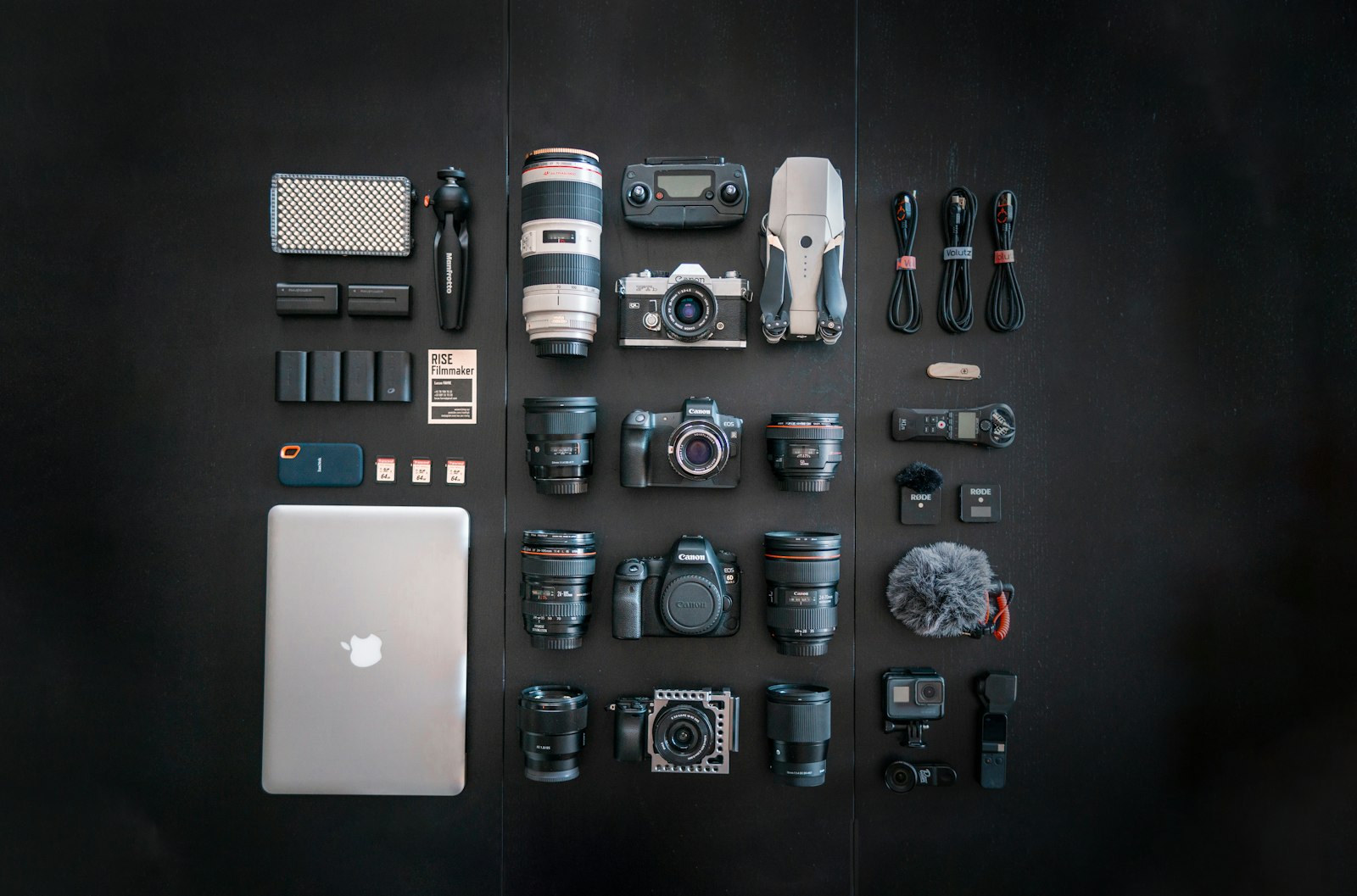 camera bodies, lenses, batteries, cables, and a laptop arranged neatly in a grid on a dark surface