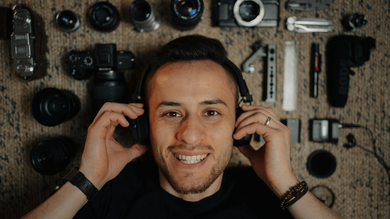 smiling man wearing headphones surrounded by camera bodies and lenses arranged on a surface
