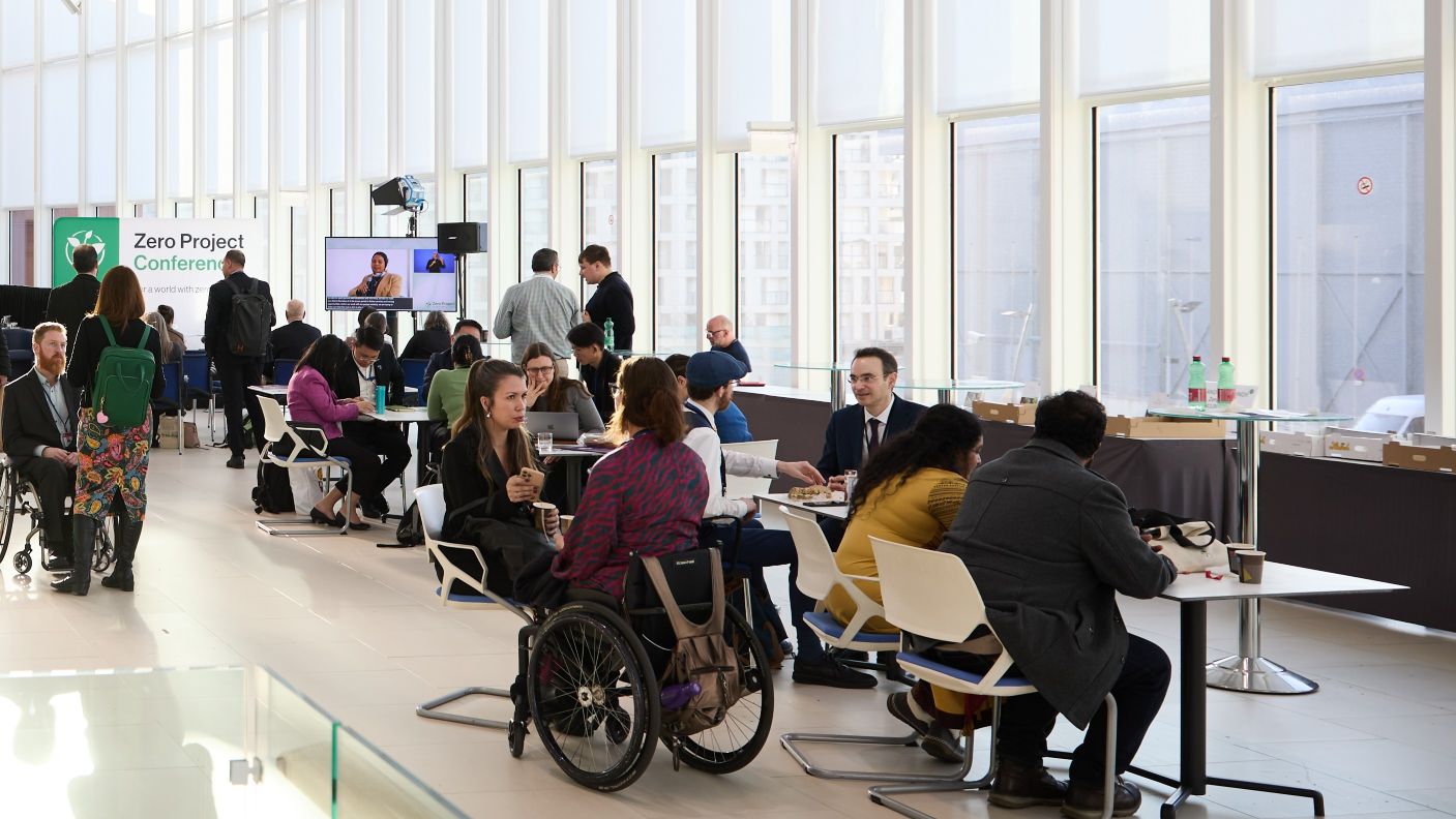 A photo of conference participants seated at coffee tables in the M-Building. Tables with water bottles and sandwiches are visible in the background