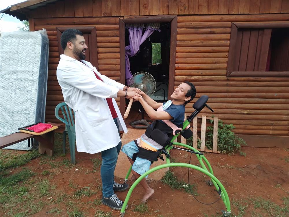 In the photo, we see a heartwarming interaction between two individuals outside a wooden structure. On the left, a man with a beard, wearing a white coat suggesting he may be a healthcare professional, is holding the hands of the person on the right, who is seated in a specialized green mobility aid, which appears to be a wheelchair. The seated individual, who seems to have a disability, is smiling joyfully during the interaction. The setting appears informal and outdoors, with personal items and a fan visible in the background, indicating a casual, possibly home or community environment. The image conveys themes of care, assistance, and the positive impact of human connection.