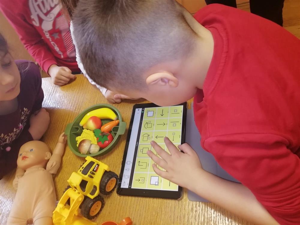 This image shows a group of young children engaged in play and learning activities. A child in a red shirt is interacting with a tablet, possibly using an educational app, while other children are around with toys like a doll and a yellow toy truck. The presence of various toys suggests a playful and inclusive environment that encourages learning through play, irrespective of gender or ability. The children appear focused and are likely developing important cognitive and social skills in a supportive setting.