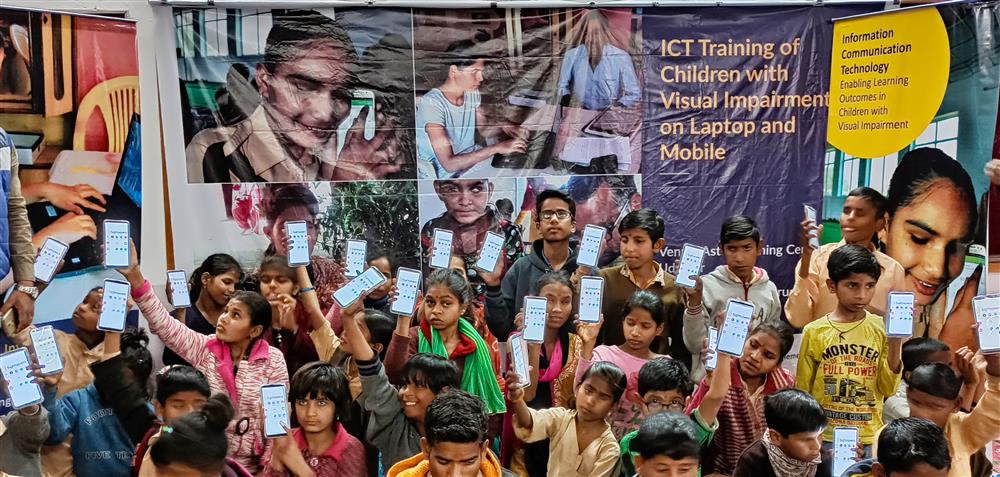 The photo shows a group of children and young adults, likely of South Asian origin, gathered in front of a banner. The banner highlights the ICT (Information and Communication Technology) training of children with visual impairment on laptops and mobiles. The children are holding up what appear to be smartphones with screens displaying an interface, possibly indicating a tool or app designed to assist visually impaired users. The image conveys a sense of community, learning, and the empowering role of technology in enhancing the lives of individuals with disabilities. The setting suggests an event or gathering to promote accessibility and inclusion through technological education.