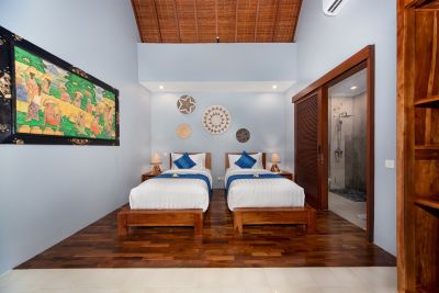 Serene Balinese twin bedroom with wooden beds, large landscape painting, woven wall decor, and sliding door to en-suite shower.