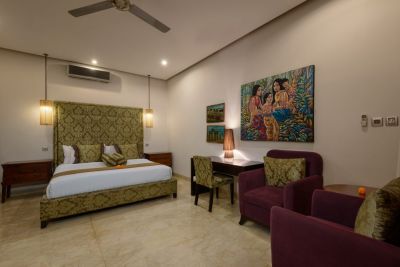 Luxurious Balinese bedroom with green damask headboard bed, purple armchairs, desk, ceiling fan, and colorful tropical artwork.