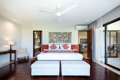 Spacious Balinese bedroom with king bed, red accents, carved wooden headboard, ceiling fan, armchair, and sliding glass doors to lush outdoor views.