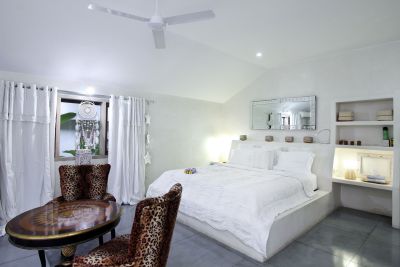Cozy tropical bedroom with white platform bed, leopard-print armchairs, dreamcatcher window, and ceiling fan.