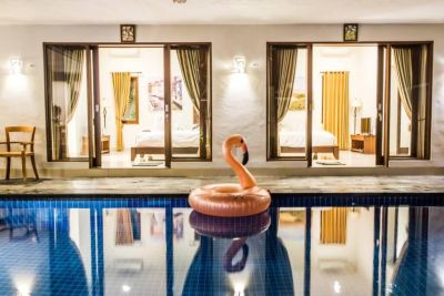 Nighttime poolside view of a luxurious tropical villa with an inflatable pink flamingo float in the blue water and lit open bedroom doors in the background.